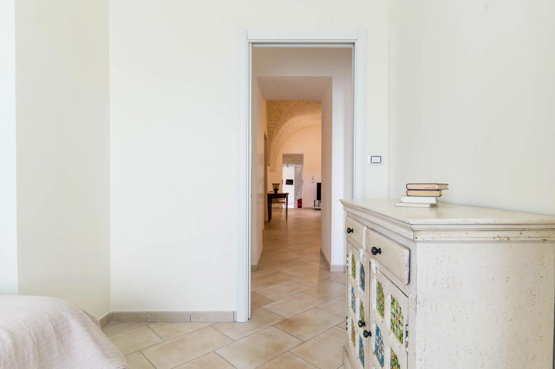 Un pasillo interior luminoso da a otra habitación, con suelo de baldosas y un mueble.
