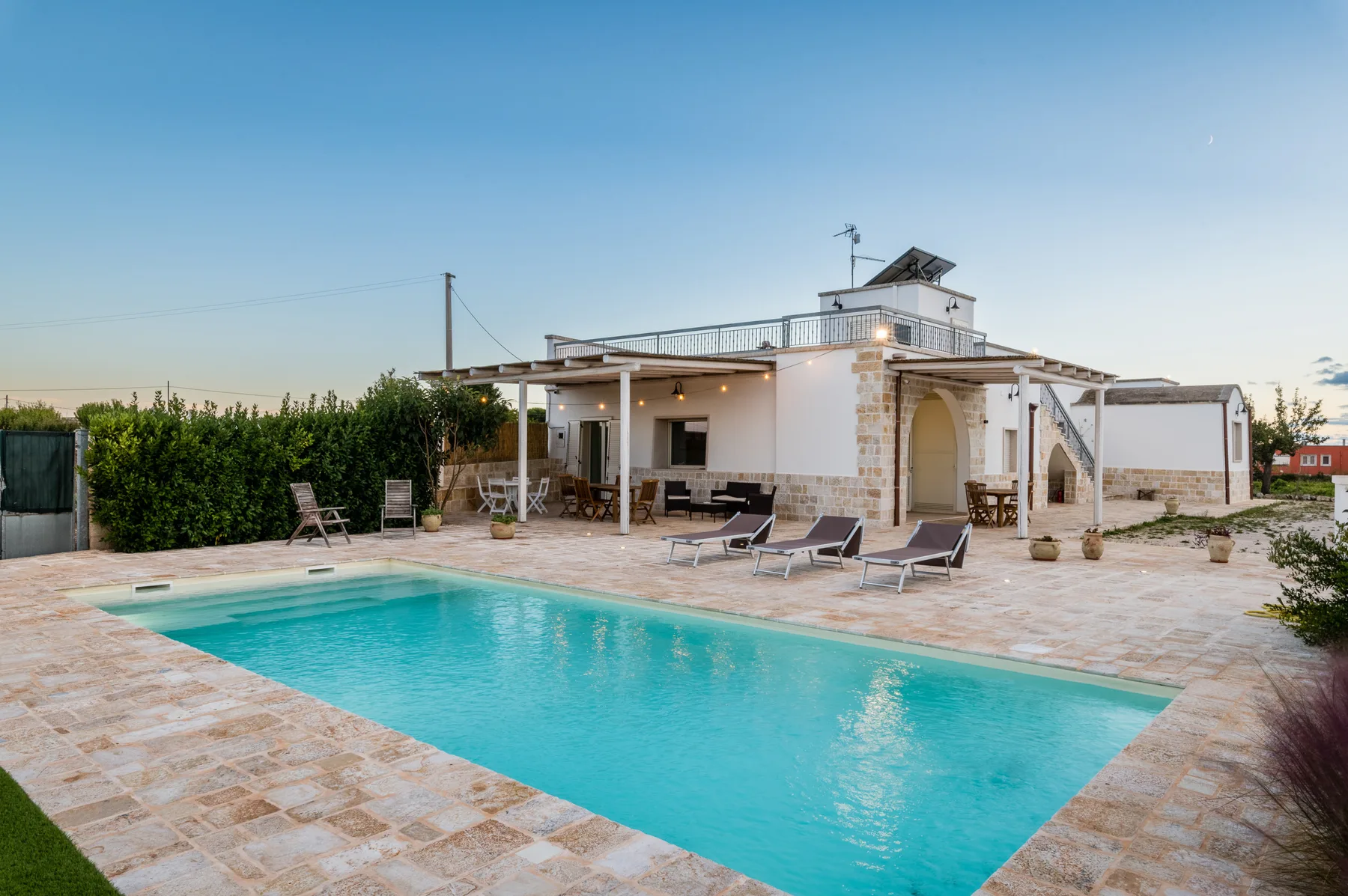 Una piscina está junto a un patio exterior blanco con asientos para descansar.