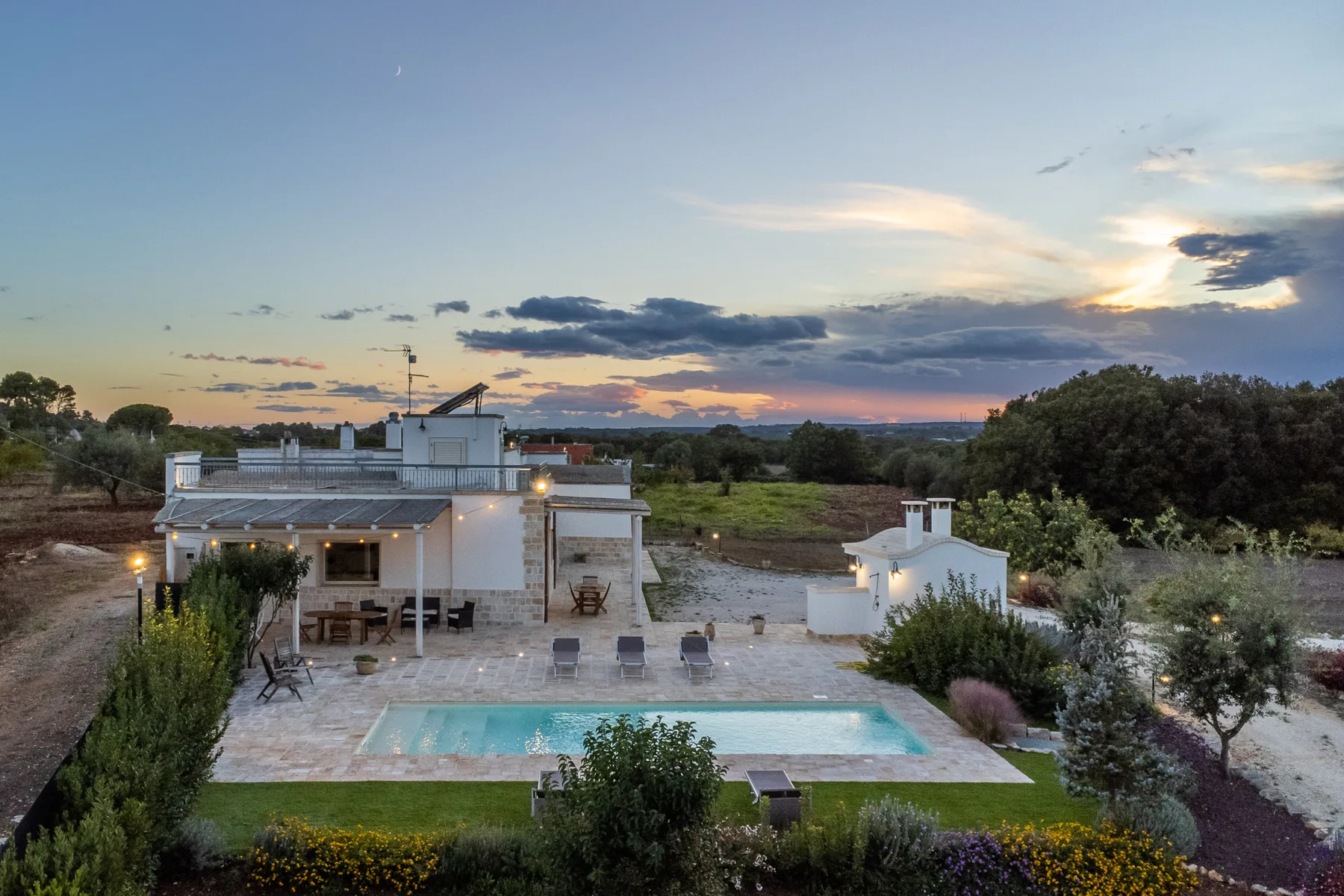 Una casa moderna y una piscina están en un jardín ajardinado al atardecer.