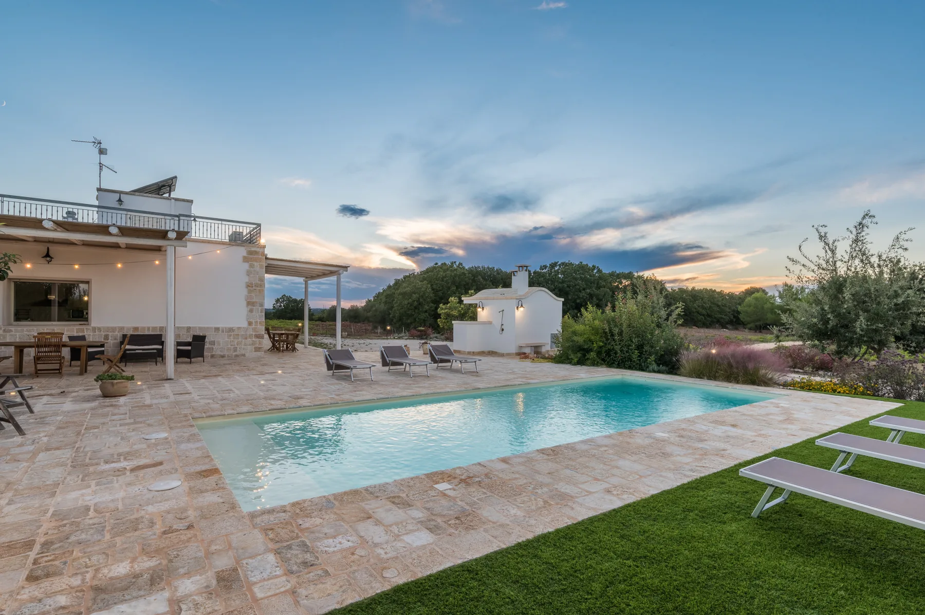 Una piscina al aire libre junto a un patio de piedra y un jardín al atardecer.