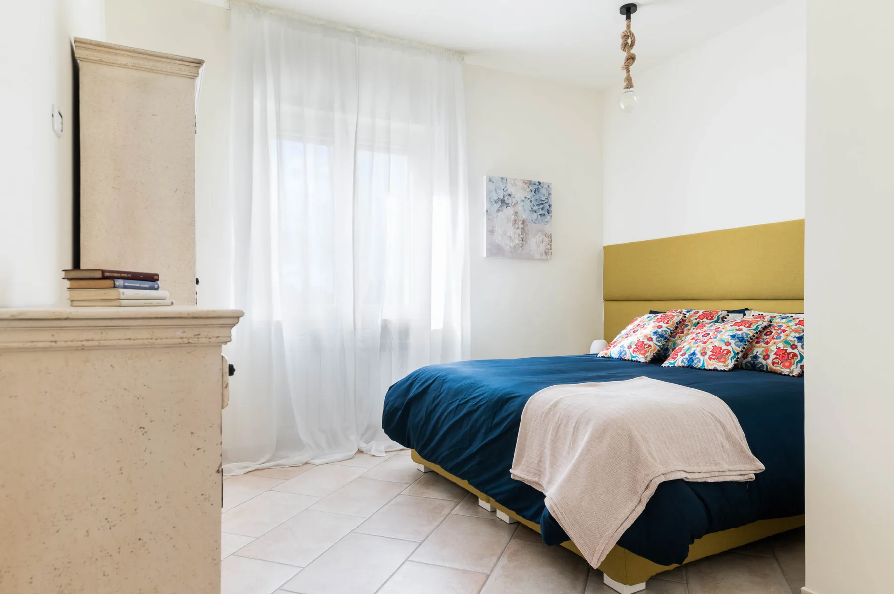 Un dormitorio con una cama vestida de azul junto a una ventana luminosa con cortinas.