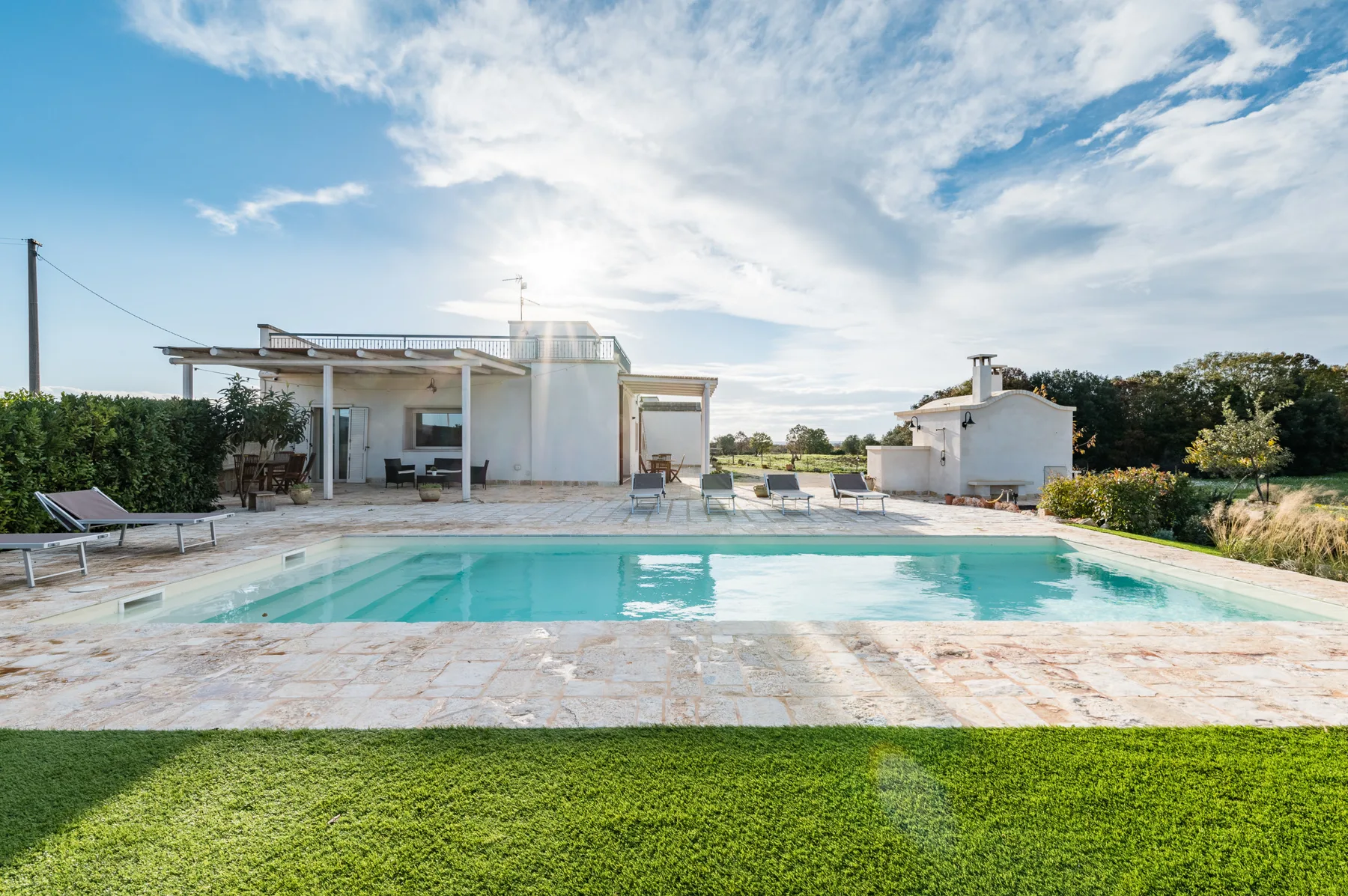 Piscina al aire libre junto a una casa blanca, con terraza pavimentada y césped.