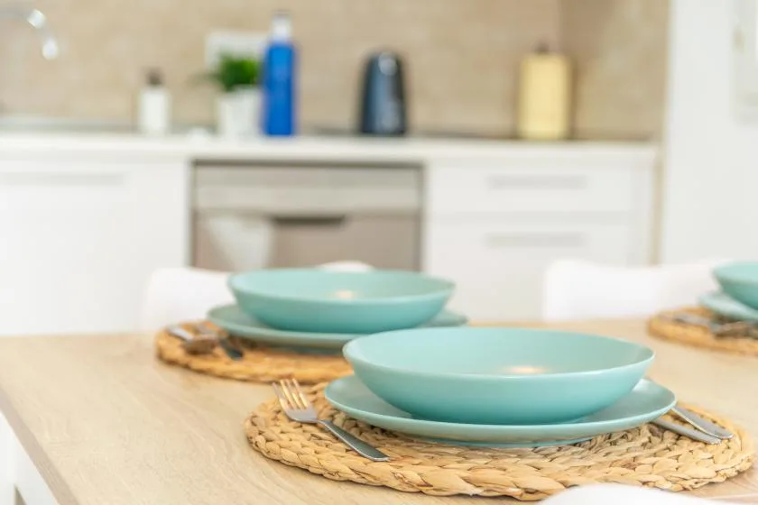 A dining table is set with turquoise dishes and woven placemats in a bright kitchen.