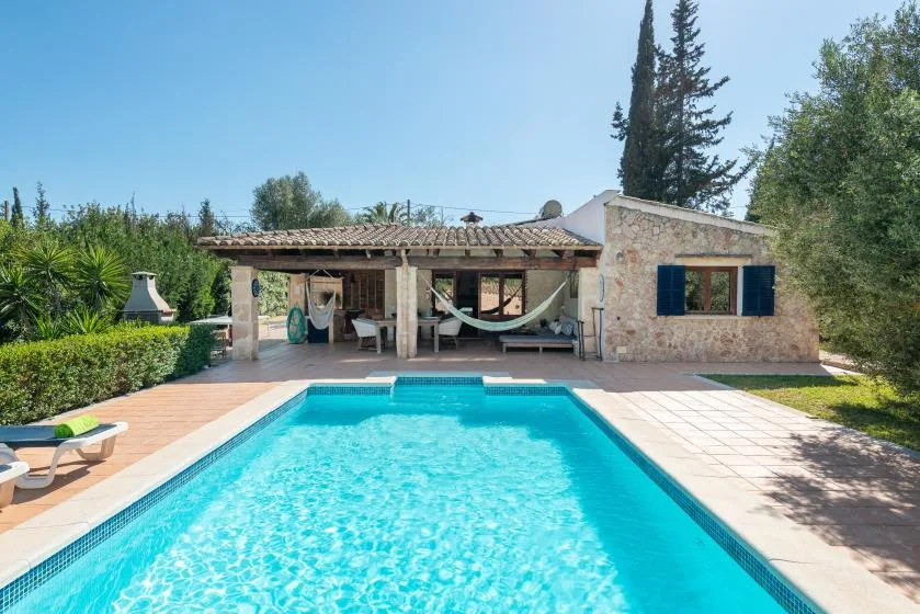 Una piscina frente a una casa de piedra con un patio sombreado.
