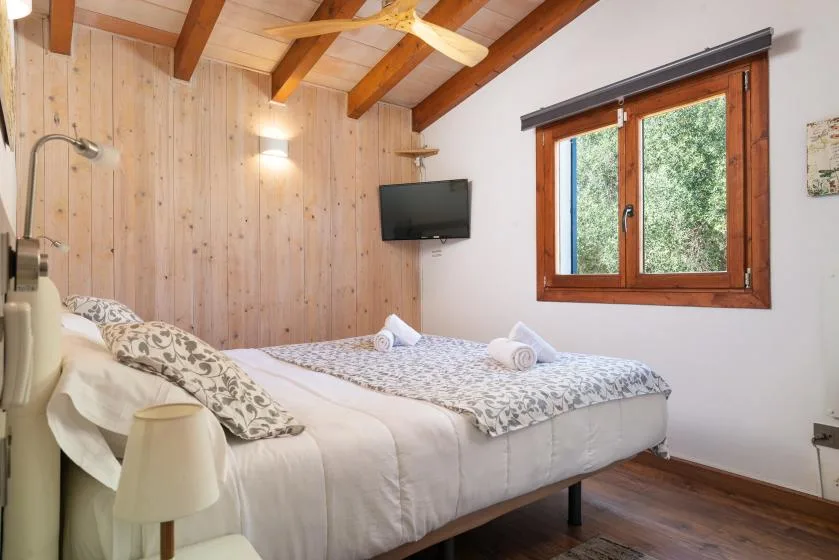 A bedroom with a double bed, wood-paneled walls, and a window overlooking greenery.
