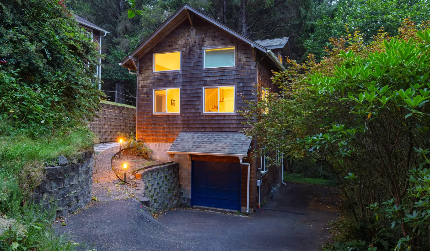 A wooden house with lit windows sits among dense greenery beside a paved drive.