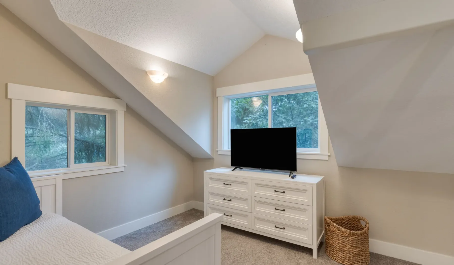 Un dormitorio luminoso con cama, cómoda y ventanas con vista a los árboles.