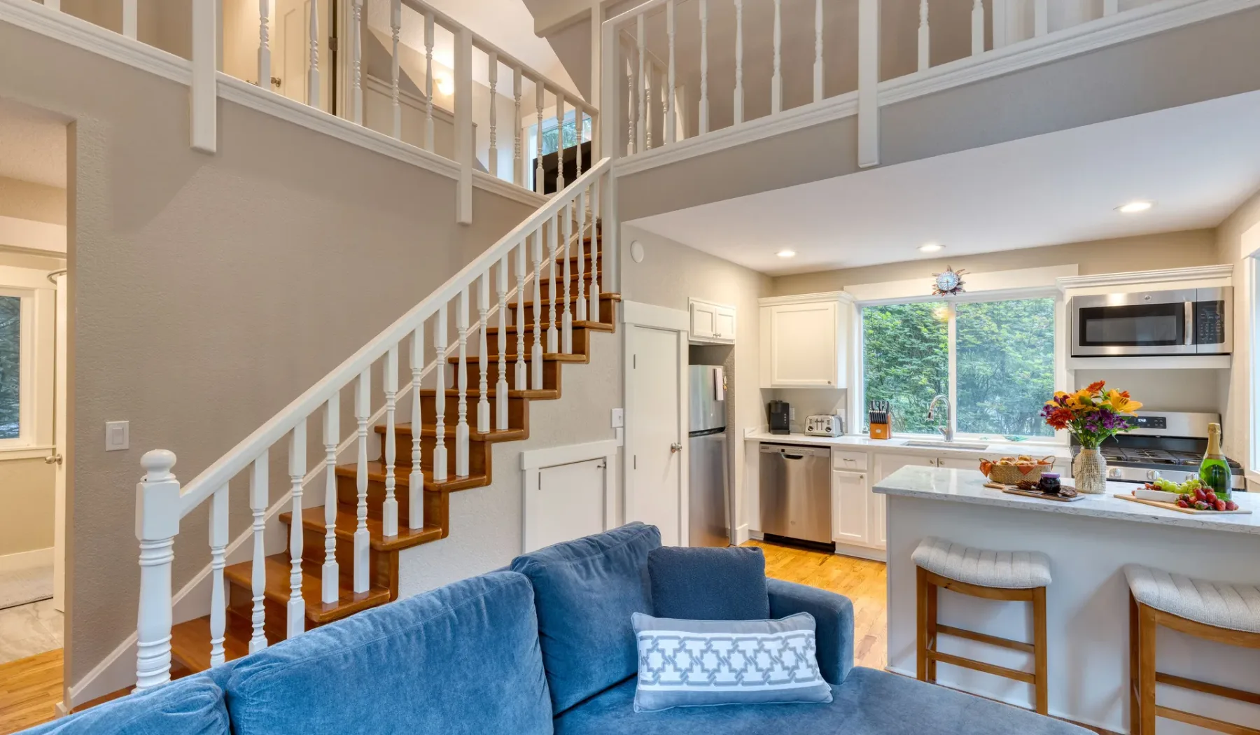 A bright kitchen and living area sits beside a staircase with white railings.