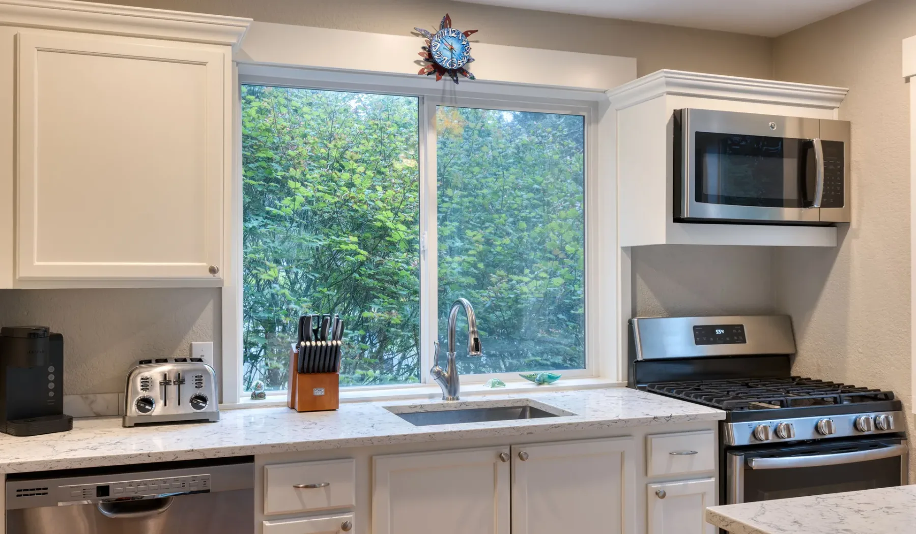 Una cocina luminosa con gabinetes blancos, un fregadero y una gran ventana con vista a la vegetación.