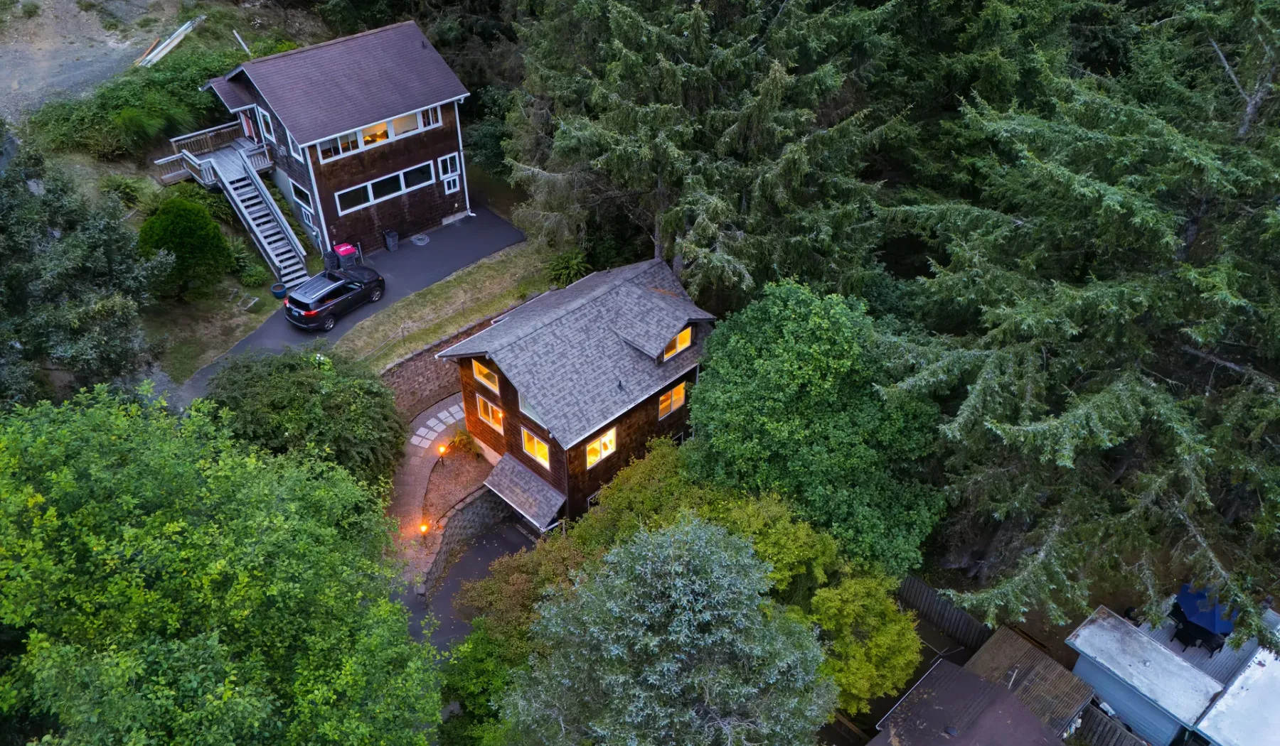 Vista aérea de dos casas rodeadas de árboles frondosos, con luces encendidas en la casa inferior.