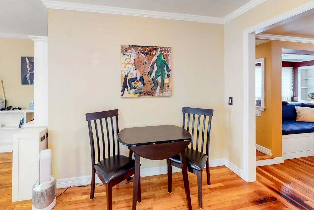 A small dining area has a wooden table and chairs beneath a colorful painting.