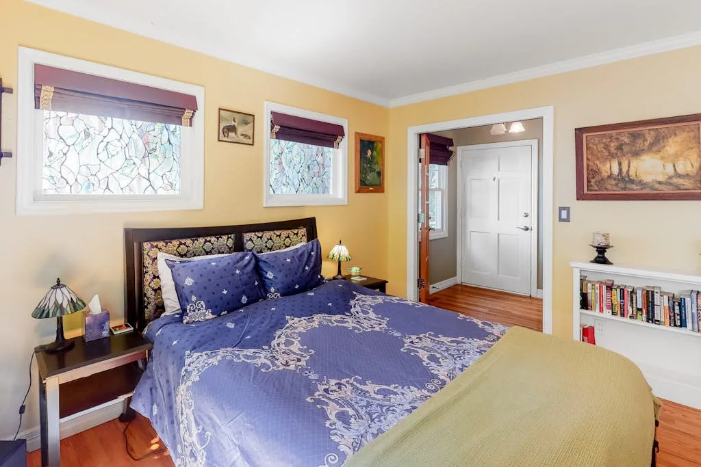 A bedroom with a bed, patterned blue bedding, and two windows.