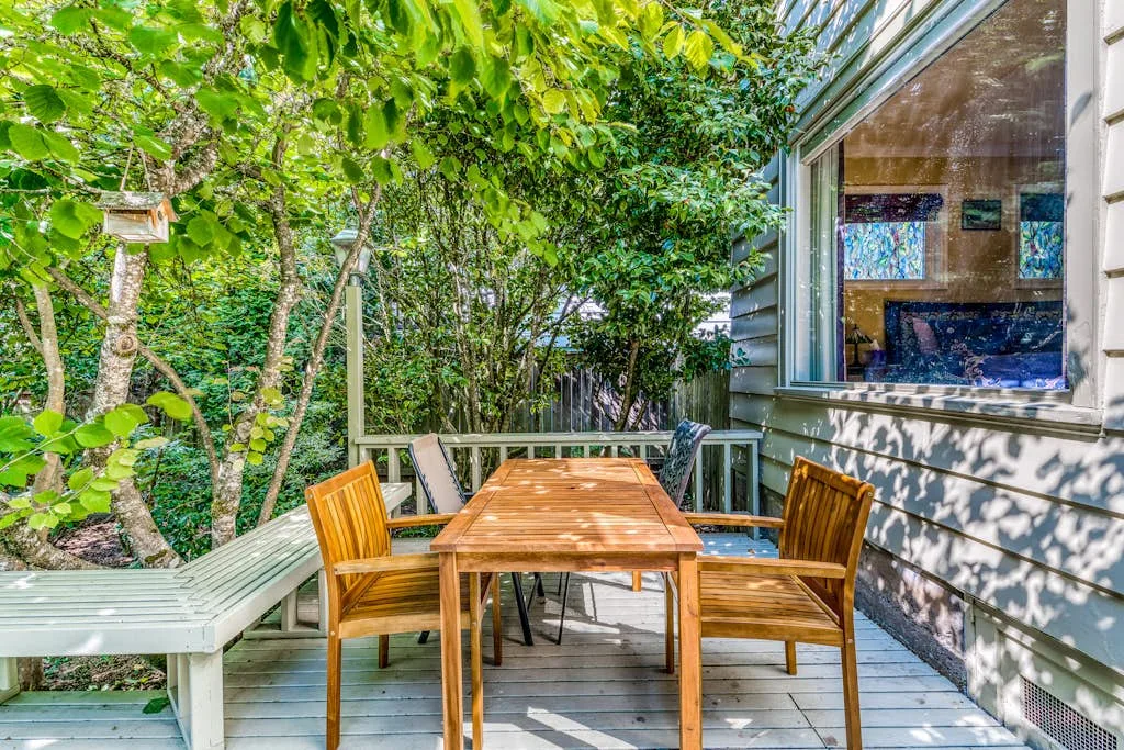 A wooden outdoor dining table and chairs sit on a leafy deck beside a window.