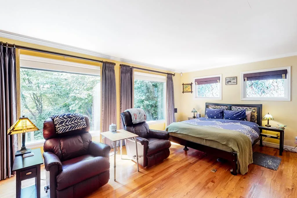 A bedroom with a bed and two armchairs beside large windows.
