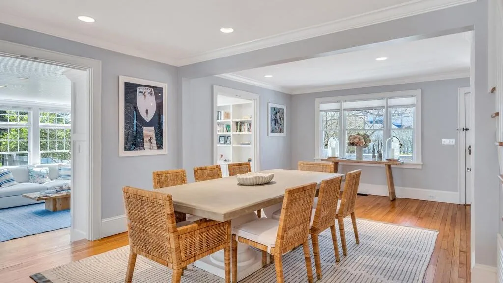 A dining area with a large table and woven chairs beside a bright sitting room.