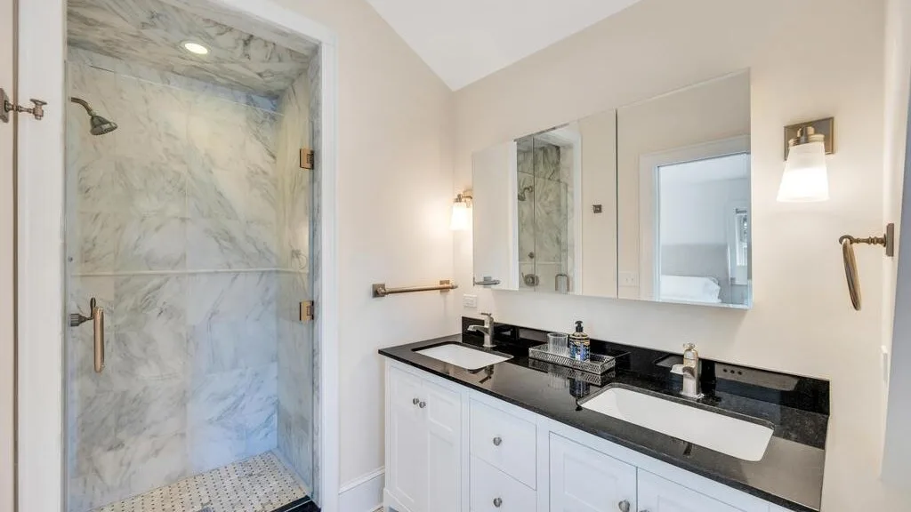 A bright bathroom with a long vanity, two sinks, and a marble shower.
