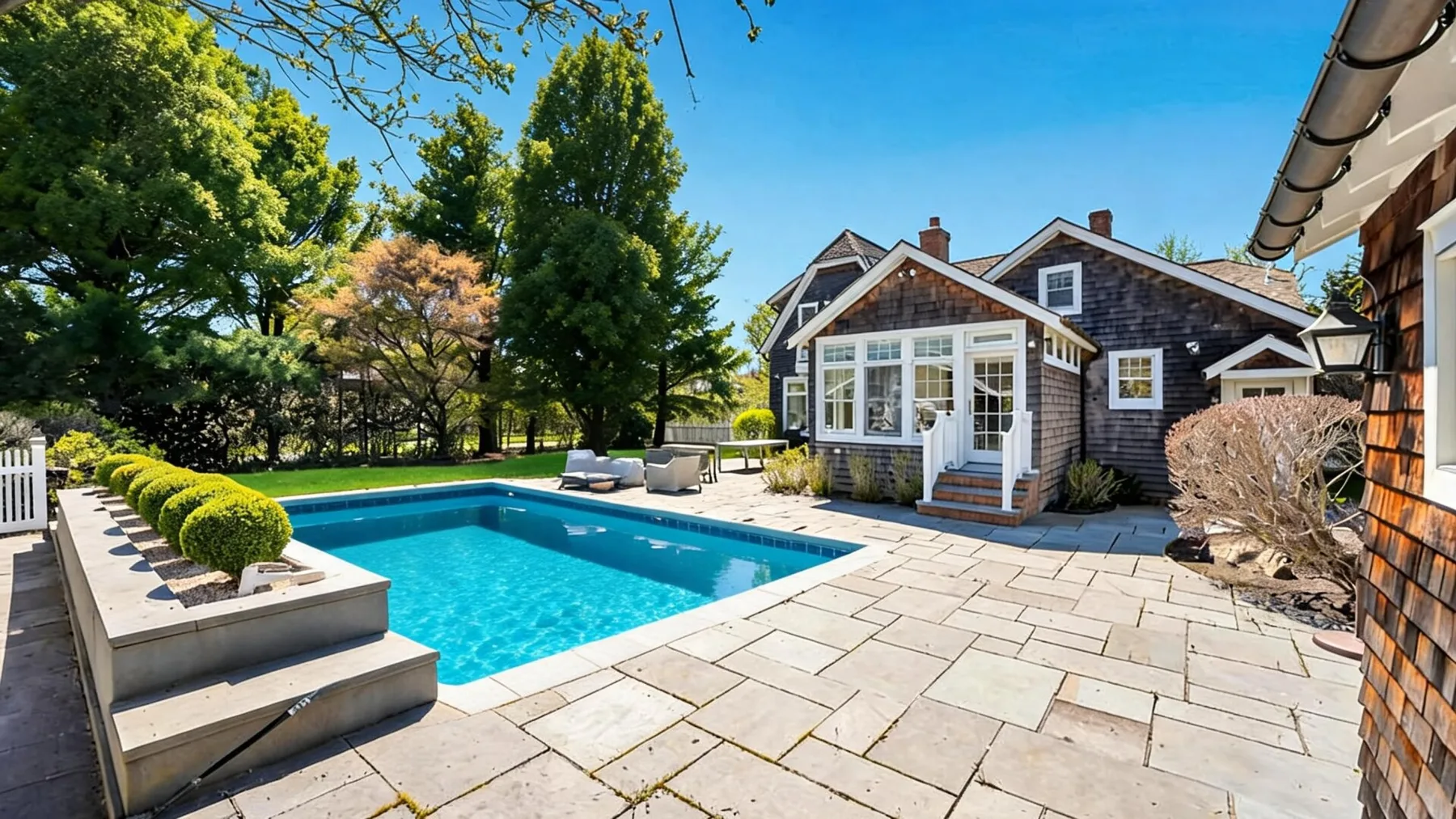 A backyard pool beside a shingled house, with a paved patio and surrounding trees.