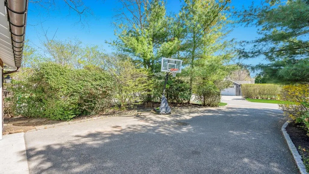 An outdoor paved basketball area with a hoop surrounded by trees and shrubs.