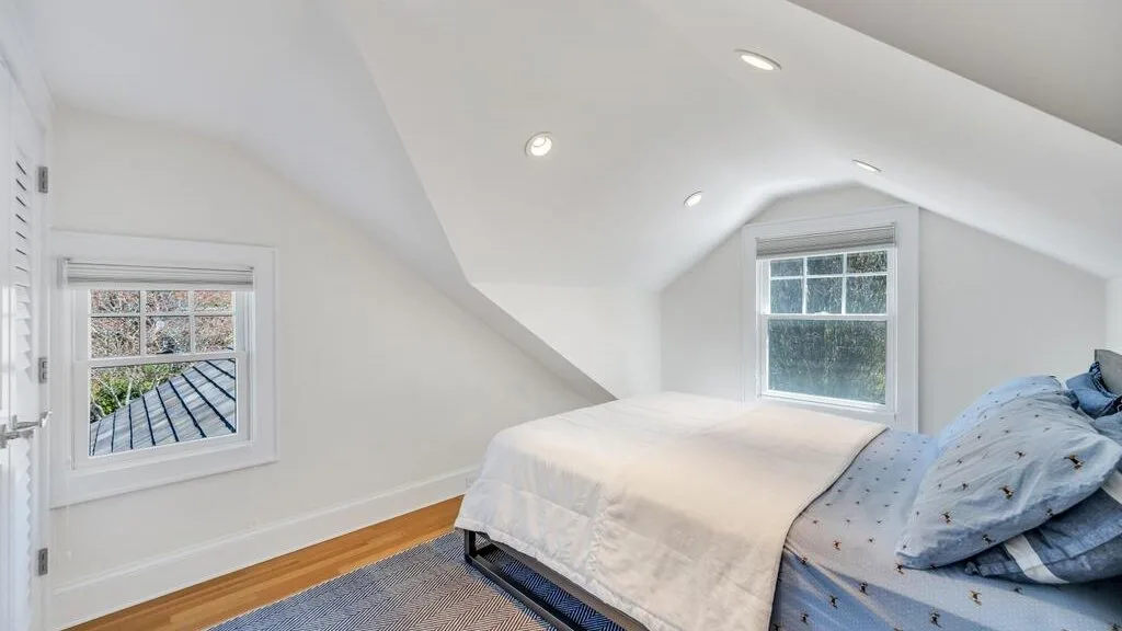 A bright attic bedroom with a bed beneath sloped ceilings and two windows.