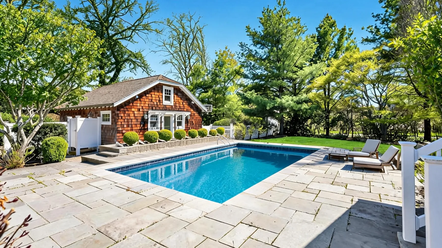 An outdoor swimming pool sits beside a paved patio and a small shingled building.