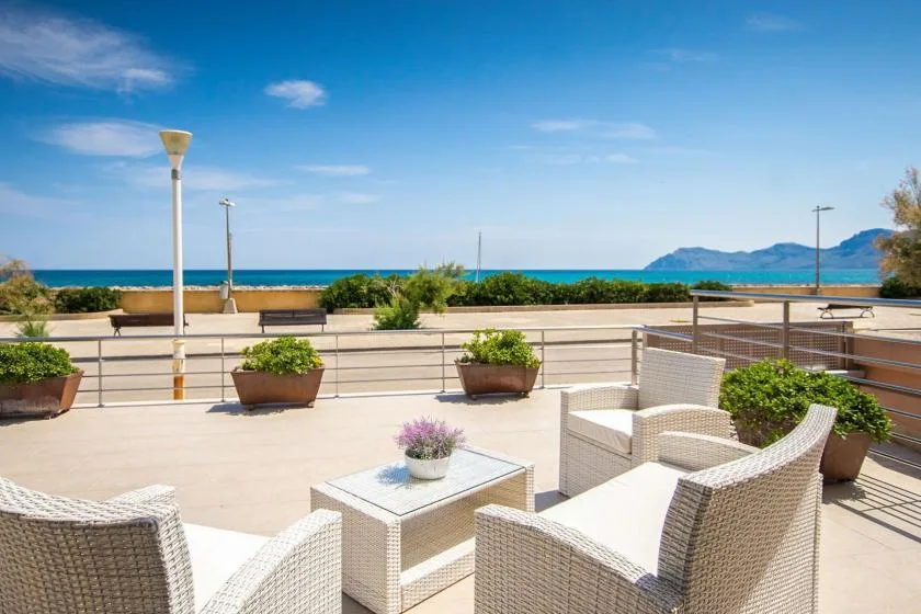 A sunny outdoor terrace with wicker seating overlooks the sea and distant mountains.