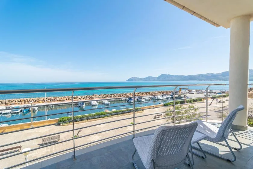 A balcony overlooks a marina and the sea, with outdoor seating in the foreground.