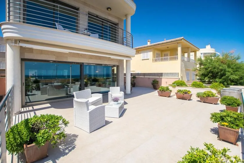A sunny outdoor terrace with seating, potted plants, and a glass-fronted building.
