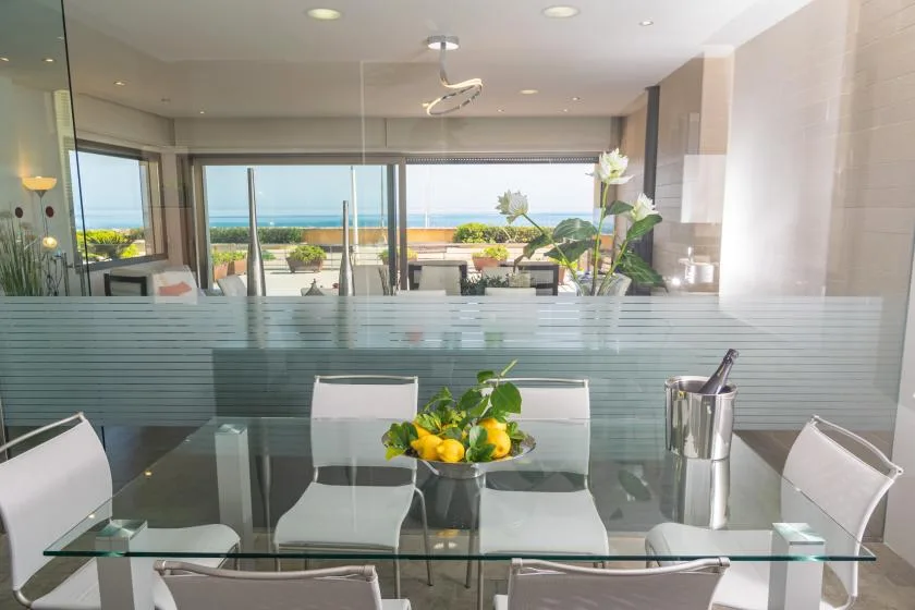 A glass dining table sits beside a frosted glass divider, with ocean views beyond.