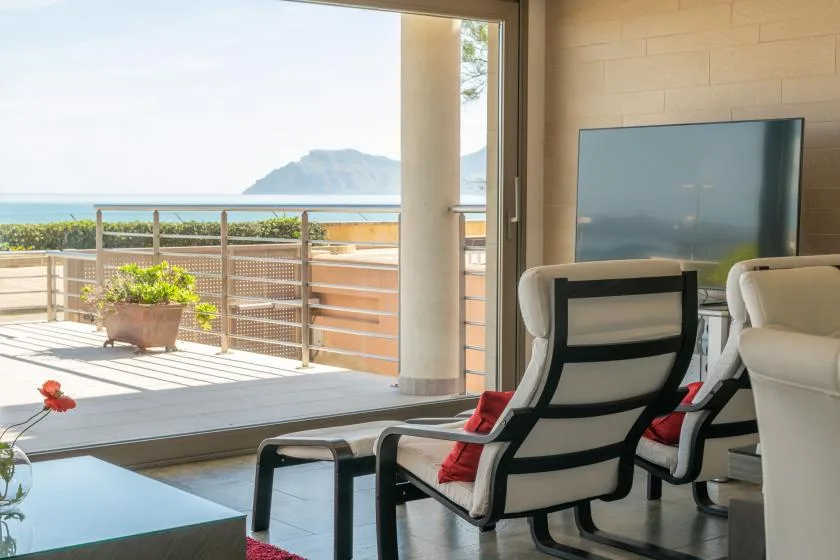 A living area opens onto a sunny terrace with seating and a view of the water.