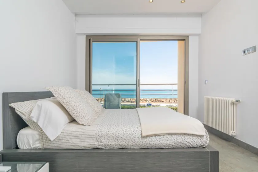 A bright bedroom with a bed beside large windows overlooking the sea.