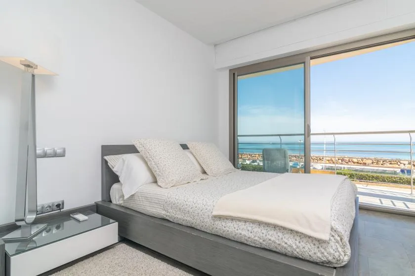 A bright bedroom with a bed beside large windows overlooking the sea.