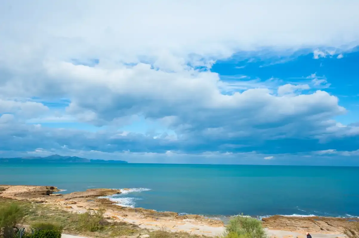 A coastal landscape shows turquoise sea beside a rocky, scrub-covered shore.