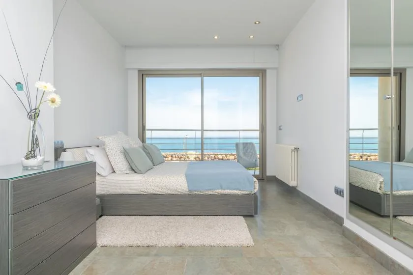 A bright bedroom with a bed facing large windows overlooking the sea.