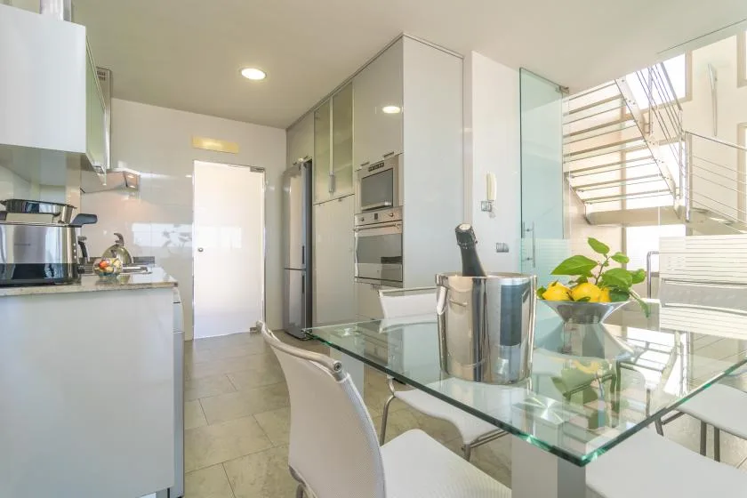 A bright modern kitchen and dining area with glass table and nearby staircase.