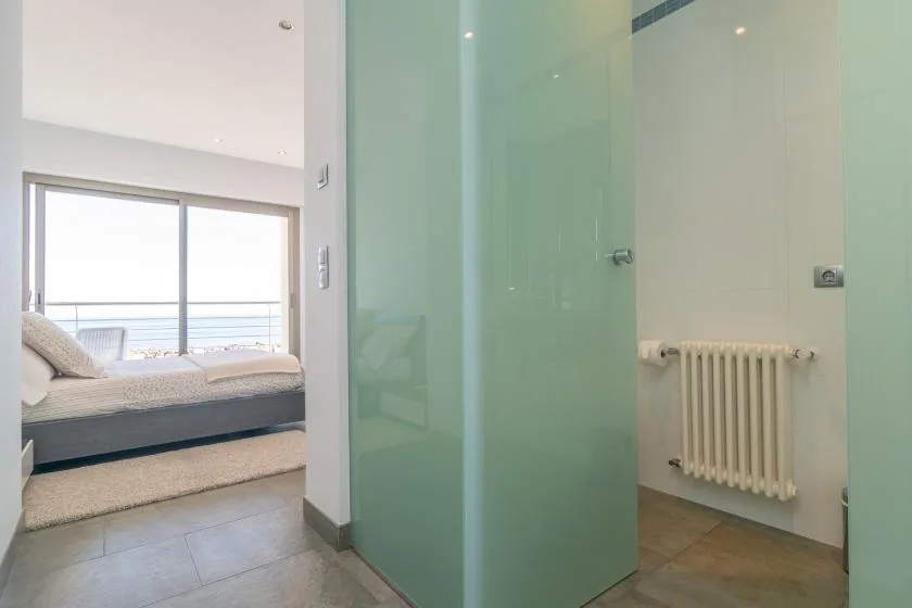 A glass shower area opens onto a bedroom with a bed beside a large window.