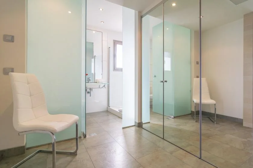 A bright bathroom with glass partitions and a pale upholstered seat.