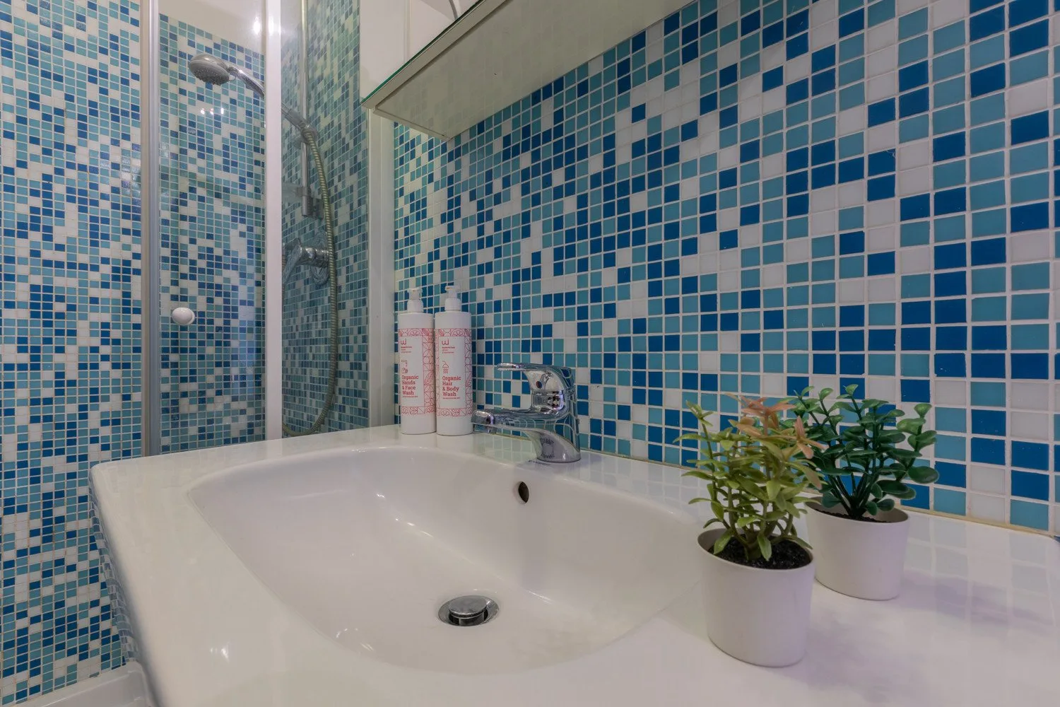 A bathroom sink sits against blue mosaic tile walls, with small potted plants nearby.