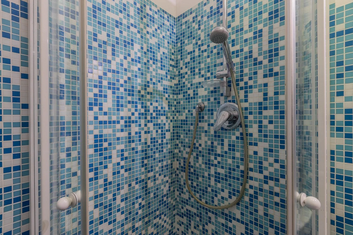 A tiled shower with a handheld showerhead and wall-mounted controls.