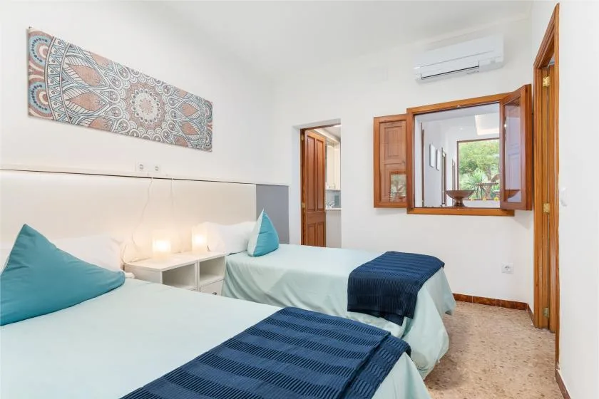 A bright bedroom with two beds, teal cushions, and a patterned wall hanging.