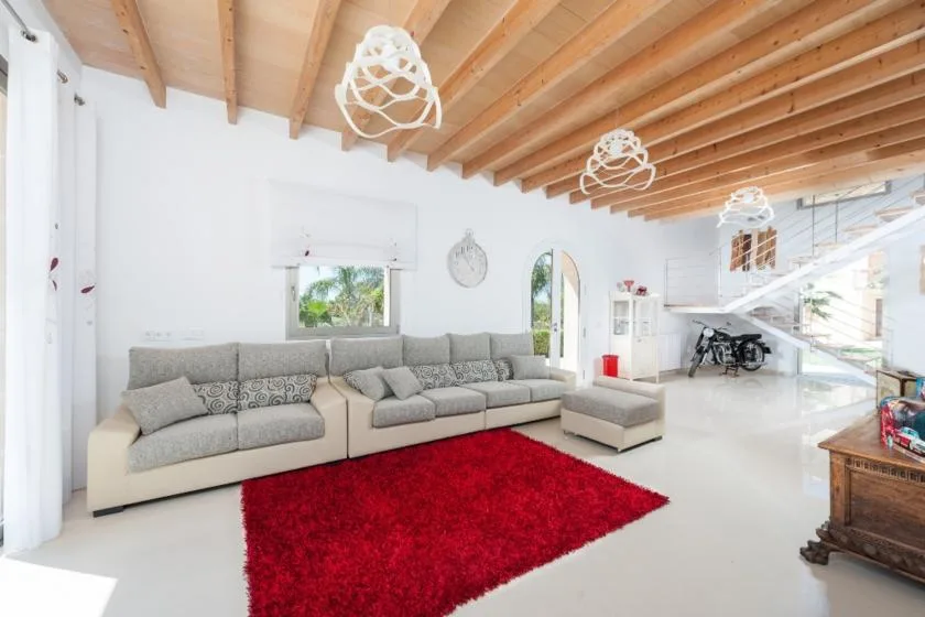 A bright living room with a sectional sofa and red rug beneath exposed wooden ceiling beams.