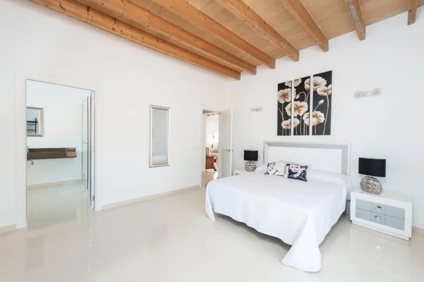 A bright bedroom with a white bed beneath exposed wooden ceiling beams.