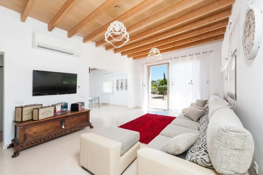 A bright living room with pale seating, a red rug, and a wooden-beam ceiling.