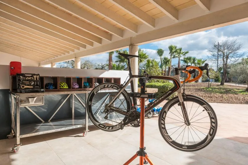 A bicycle stands on a repair stand beside a workbench in a covered outdoor workspace.