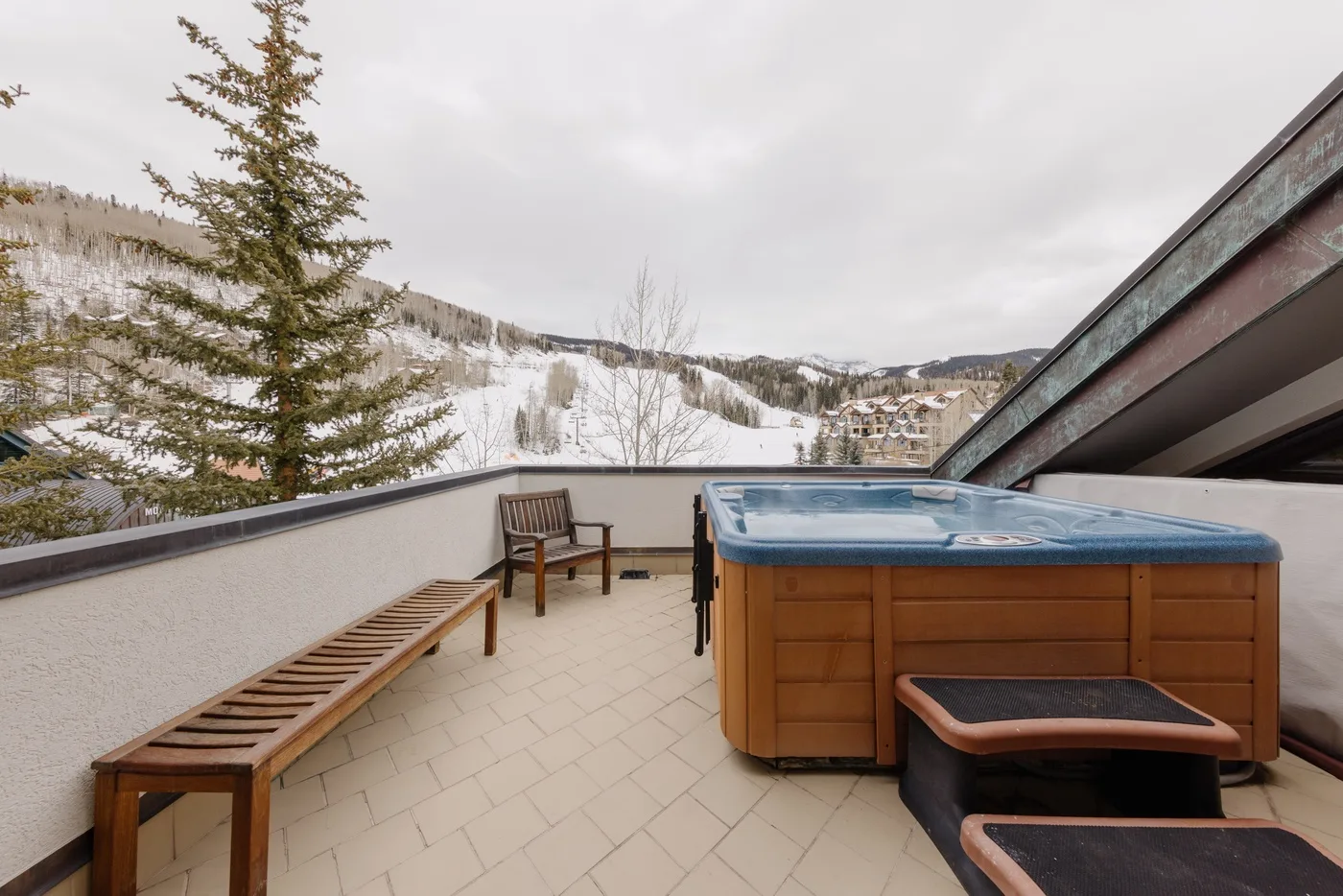 A rooftop terrace overlooks a snowy mountain landscape, with a hot tub beside the seating.