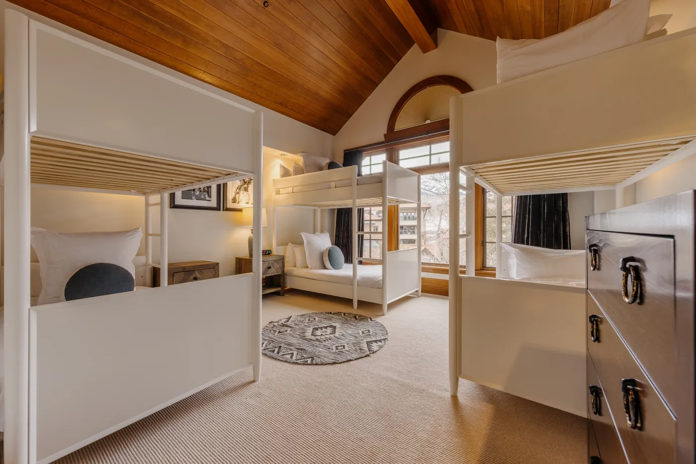 A bunk bedroom with white beds, a round rug, and a sloped wood ceiling.