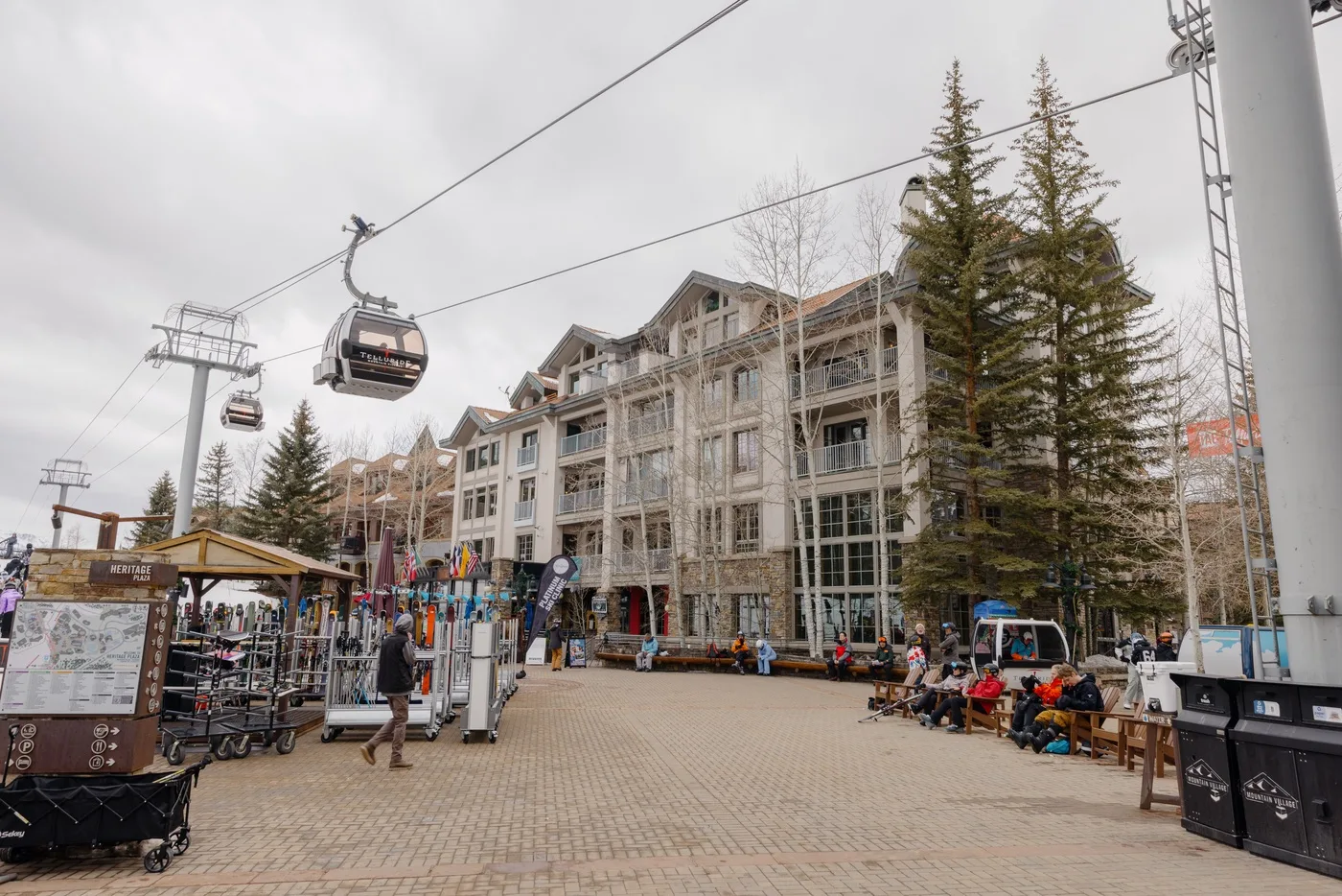 A paved plaza with a large building, outdoor seating, and an overhead cable car.
