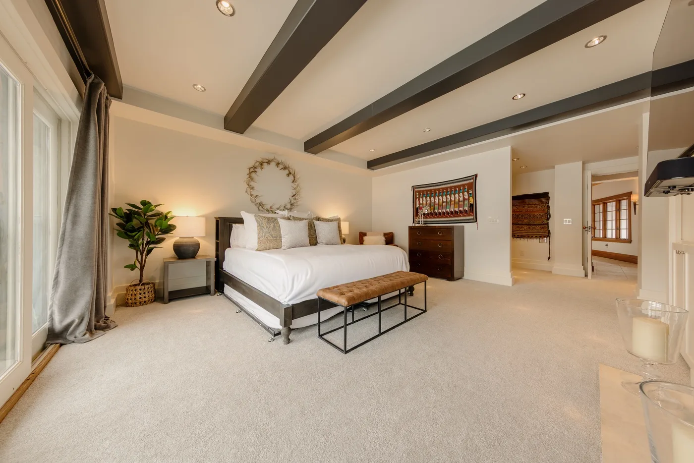 A bright bedroom with a large bed, a bench, and dark ceiling beams.