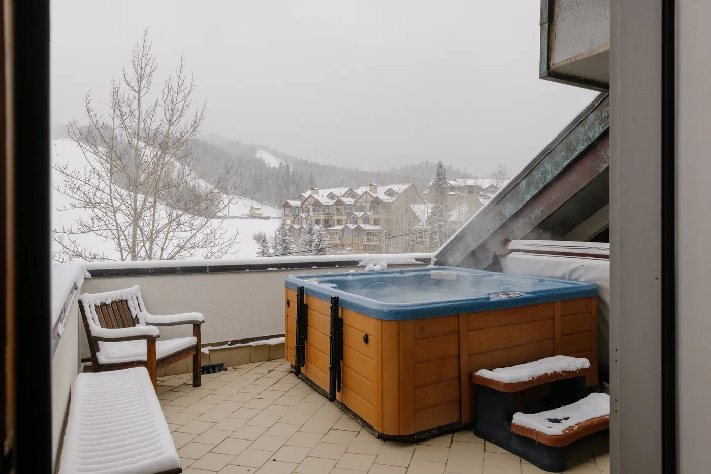 A snowy outdoor terrace features a steaming hot tub and a view of wooded hills.