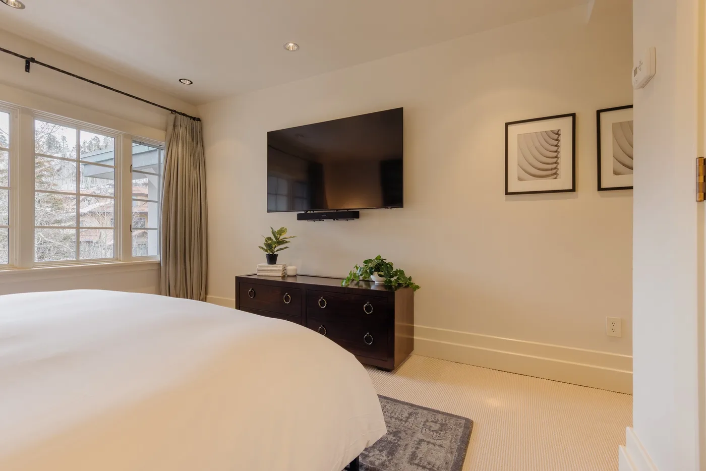 A bedroom with a bed, wall-mounted television, and dresser beside a window.