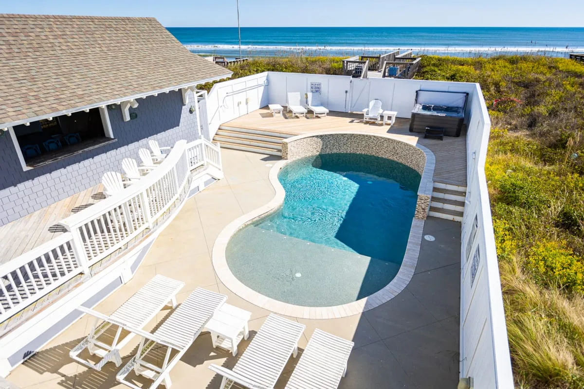 Una piscina exterior y su terraza están junto a una casa, con el océano al fondo.